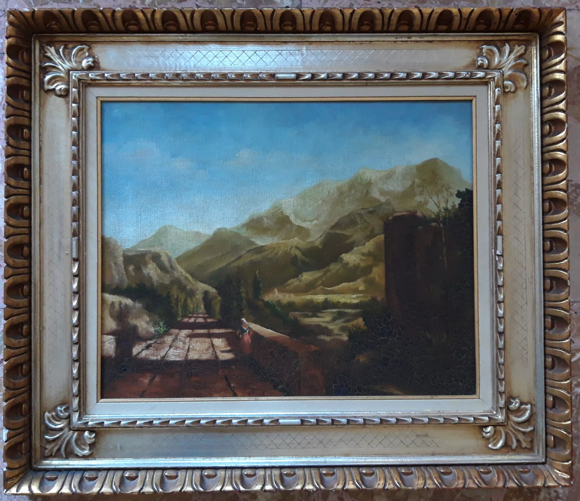 Mountain-landscape-with-intentional-craquelure-Aly-Mohsen "Painting by Aly Mohsen, executed in Oil on canvas (brush). The title is Mountain landscape with intentional craquelure and it belongs to the category Landscapes. In Mountain landscape with craquelure, Aly Mohsen presents a wide and silent vision of the mountain environment, where a paved road guides the viewer’s gaze toward an horizon of hills and mountains immersed in soft, diffused light. A small and discreet human presence establishes a sense of scale and reinforces the dialogue between humankind and nature, suggesting an idea of passage and contemplation. The composition is built upon strong perspectival depth, articulated through successive planes that unfold naturally toward the distant peaks. Warm earth tones and muted greens of the mountain slopes gradually blend with the blue of the sky, creating an atmosphere of suspended calm and natural balance. The brushwork is measured and narrative, capable of conveying the solidity of the ground, the softness of the hills, and the vastness of space. Deliberately, Aly Mohsen has recreated a controlled pictorial craquelure, giving the surface of the painting the appearance of an aged work. This choice is not merely aesthetic, but demonstrates the artist’s technical mastery and his ability to consciously engage with historical painting processes, particularly those of classical European landscape tradition. Light is soft and evenly distributed, guiding the viewer along the visual path and encouraging a slow, contemplative reading of the image. The overall effect is one of temporal suspension and harmony between nature and human presence. The richly decorated gilded frame further enhances the painting, reinforcing its classical character and accompanying the composition with balance and elegance. Mountain landscape with craquelure fits within Aly Mohsen’s landscape production as a work of strong formal balance and visual depth, capable of uniting historical memory, technical expertise, and poetic sensitivity."