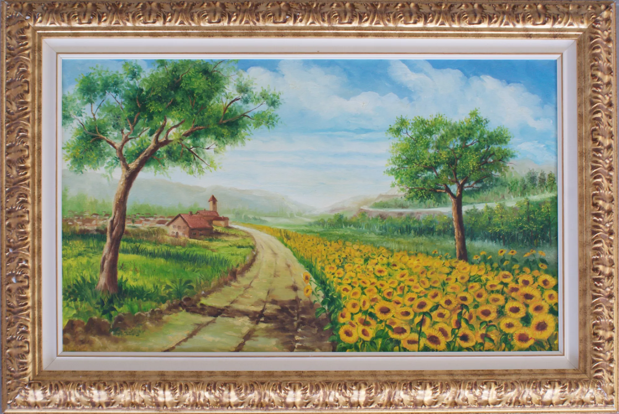 The-sunflower-path-Aly-Mohsen "Painting by Aly Mohsen, executed in Oil on canvas (brush). The title is The sunflower path and it belongs to the category Landscapes. In this painting, Aly Mohsen depicts a luminous and welcoming rural landscape, rooted in the figurative tradition of European and Mediterranean landscape painting. A path crosses the composition and guides the viewer’s gaze toward a small village set within greenery, establishing a spatial structure based on movement and visual continuity. The road, as the central compositional element, functions as a visual metaphor for journey and connection between nature and human presence. On the right, a wide field of sunflowers in full bloom illuminates the landscape with warm, vibrant tones, while on the left leafy trees and green meadows balance the composition with a sense of freshness and stability. The clear sky, crossed by light clouds, contributes to a serene and contemplative atmosphere, free from dramatic emphasis. The soft brushwork and carefully modulated light create depth and natural coherence, highlighting the artist’s direct relationship with landscape and natural rhythms. The painting is enhanced by a richly decorated, classically styled gilded frame, which encloses the work with elegance and strengthens the dialogue between the painted surface and its support, emphasizing its formal and decorative presence."