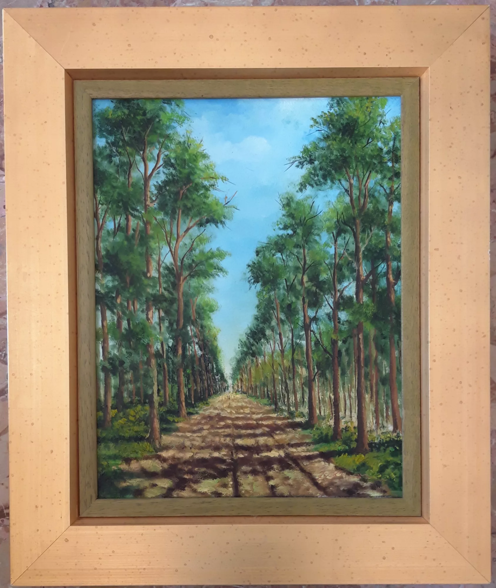 The-avenue-in-the-woods-Aly-Mohsen "Painting by Aly Mohsen, executed in Oil on canvas (brush). The title is The avenue in the woods and it belongs to the category Landscapes. In this painting, Aly Mohsen presents a woodland landscape with a strong perspectival structure, built around a long dirt avenue extending between two orderly rows of trees. The central path guides the viewer’s gaze toward the horizon, creating a clear sense of depth and spatial continuity that evokes the idea of walking and contemplation within nature. The trees, tall and rich in green foliage, form a natural architectural framework that encloses the bright, open blue sky. The shadows cast across the ground, rendered with soft and controlled brushwork, establish visual rhythm and enhance the atmosphere of calm and balance. The chromatic palette, dominated by deep greens and warm earth tones, conveys a landscape that is vibrant yet restrained, consistent with the artist’s ongoing exploration of the relationship between light, space, and the natural environment. The work is completed by a frame in warm, delicate tones, which harmonizes with the colors of the painting and enhances its formal balance without overpowering the image."