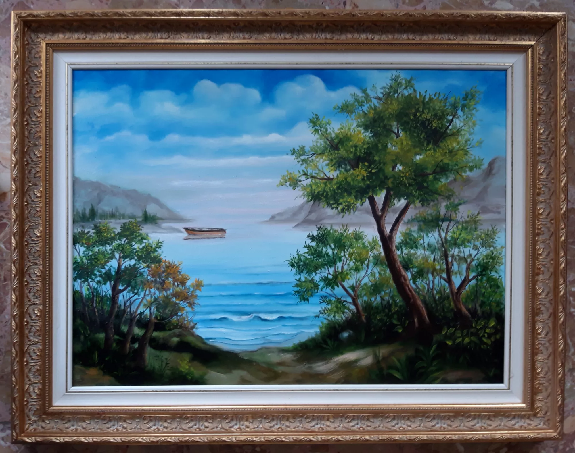 Lake-view-Aly-Mohsen "Painting by Aly Mohsen, executed in Oil on canvas (brush). The title is Lake view and it belongs to the category Landscapes. In this painting, Aly Mohsen presents a lakeside landscape characterized by calm, luminosity, and a contemplative atmosphere, where the natural environment becomes a space of silence and reflection. The composition opens onto a wide expanse of still water, marked by gentle ripples that reflect the blue sky and softly layered clouds above. In the foreground, lush vegetation and trees with lightly moving branches frame the scene, guiding the viewer’s gaze toward the center of the composition, where a small solitary boat rests upon the lake. In the background, faintly veiled mountain forms emerge with restraint, creating spatial depth and a smooth transition toward the horizon. The chromatic palette is dominated by clear blues, natural greens, and soft tonal variations, carefully balanced to convey a sense of calm and visual harmony. The brushwork is fluid and measured, effectively capturing the lightness of the air and the stillness of the water’s surface. A classically styled gilded frame completes the work, enhancing its luminosity and compositional clarity while maintaining a harmonious dialogue with the painted surface."