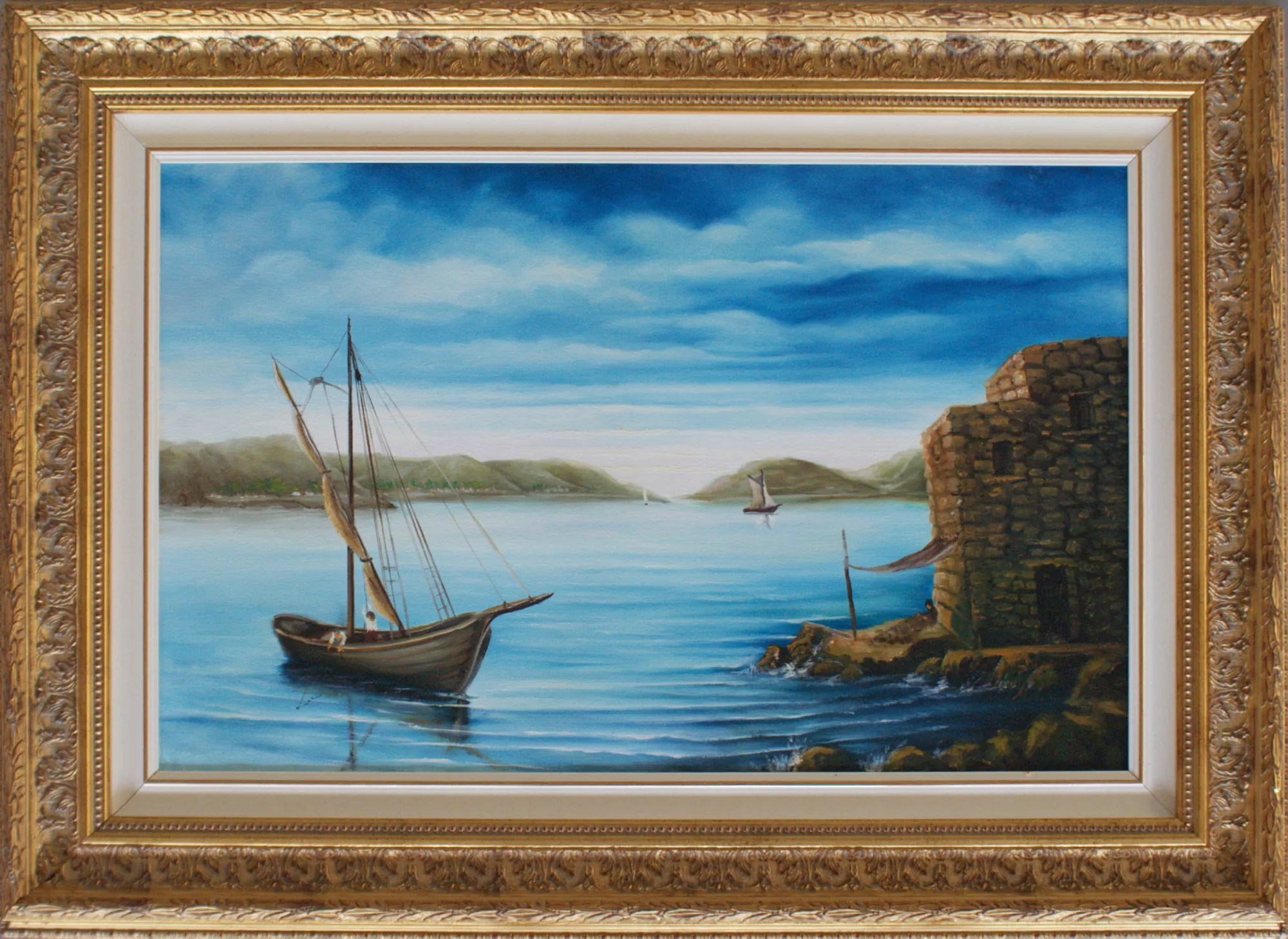 Seascape-with-stone-house-Aly-Mohsen "Painting by Aly Mohsen, executed in Oil on canvas (brush). The title is Seascape with stone house and it belongs to the category The Sea. Seascape with stone house is a painting that conveys an atmosphere of calm and contemplation, closely associated with Mediterranean coastal villages. In the foreground, a sailboat rests on still waters, reflecting the light of the sky and creating subtle chromatic effects across the surface of the sea. The light sails and slender mast introduce formal balance and vertical structure within the composition. On the right, a solid stone house overlooking the water introduces an architectural presence that dialogues with the surrounding landscape, evoking a sense of coastal life rooted in simplicity and place. In the background, gently sloping hills guide the viewer’s gaze toward the horizon, where a second boat enhances spatial depth and reinforces the perception of distance. The wide and luminous sky is rendered through delicate tonal transitions, ranging from pale blue to deeper hues, strengthening the overall sense of serenity and atmospheric continuity. The ornate, classically styled gilded frame encloses the painting with refinement, enhancing its luminosity and reinforcing its timeless visual character."