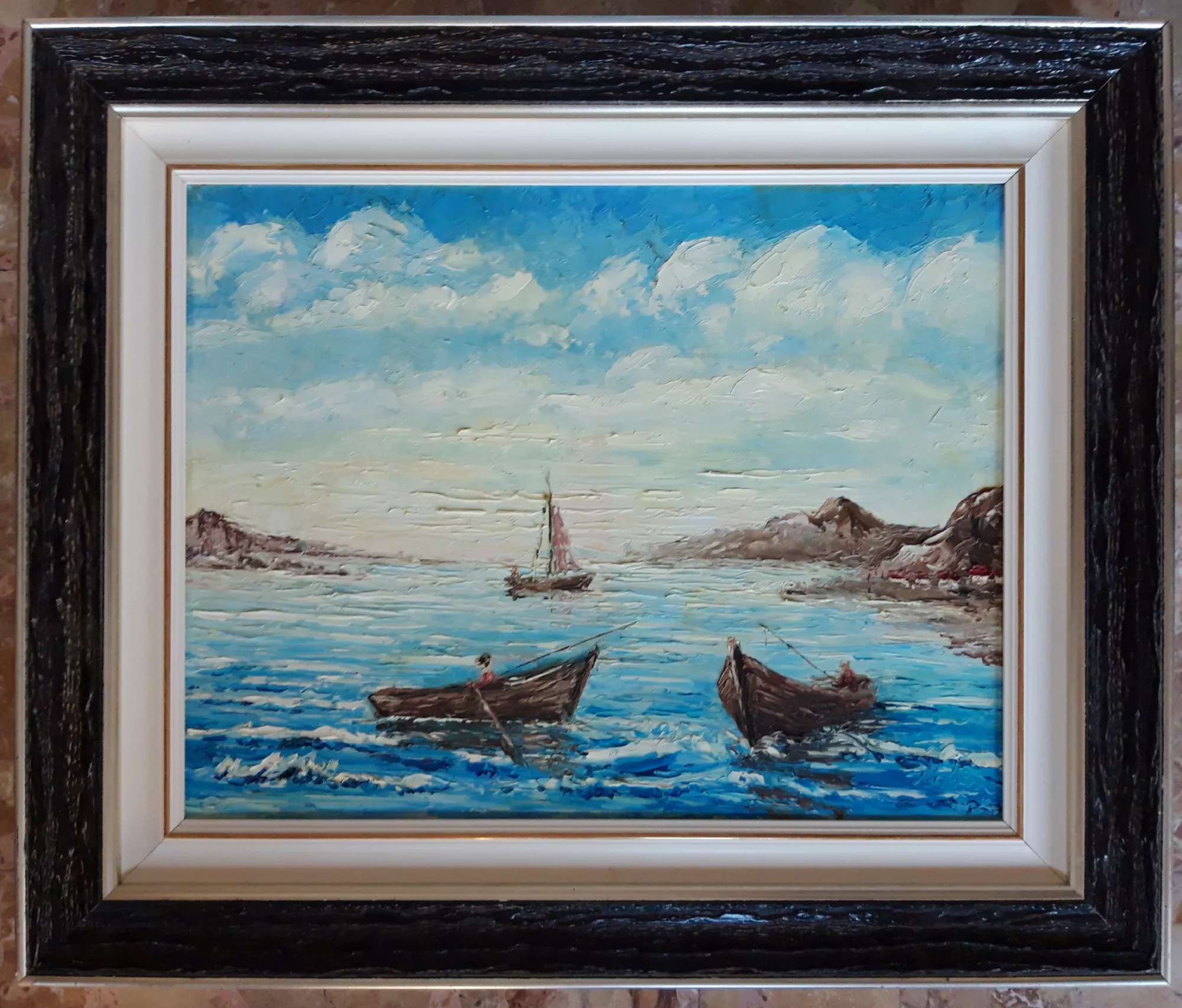 Seascape-with-boats-Aly-Mohsen "Painting by Aly Mohsen, executed in Oil on canvas (palette knife). The title is Seascape with boats and it belongs to the category The Sea. Seascape with boats is a palette-knife painting in which Aly Mohsen explores life at sea through a richly textured and dynamic pictorial language. In the foreground, two small wooden boats move across rippling waters, shaped by thick layers of paint that convey movement and depth across the marine surface. The lightly suggested figures on board evoke an everyday coastal scene marked by work, waiting, and silence. At the center of the composition, a more distant sailboat guides the viewer’s gaze toward the horizon, establishing a balanced visual relationship between foreground and background. The rocky coastlines framing the scene reinforce the compositional structure and visually open the space toward the open sea, expanding the sense of depth. The wide, luminous sky is built through layered applications of blues and whites, emphasizing air, light, and atmospheric depth. The frame, darker on the outer edge and lighter on the inner profile, enhances the chromatic contrasts of the painting and highlights the material intensity of the pictorial surface, contributing to the overall harmony of the work."