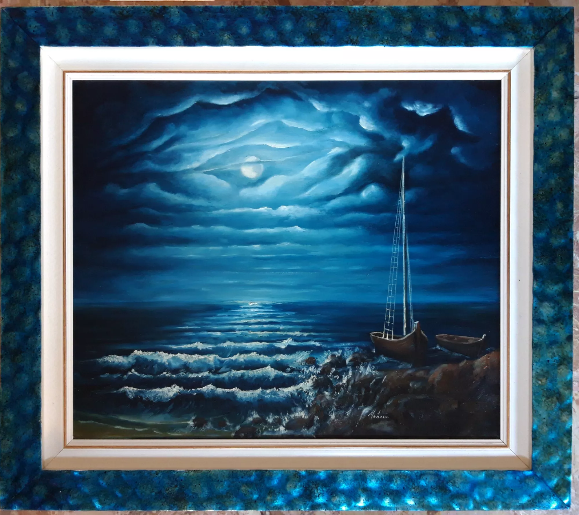 Night-seascape-Aly-Mohsen "Painting by Aly Mohsen, executed in Oil on canvas (brush). The title is Night seascape and it belongs to the category The Sea. Original work by Aly Mohsen depicting a nocturnal seascape characterized by an intense and contemplative atmosphere. The scene portrays the sea illuminated by moonlight, with waves breaking along the shore and a solitary boat in the foreground, introducing a quiet and discreet human presence within a natural setting dominated by the vastness of the horizon. The subject aligns with the tradition of Mediterranean seascape painting, reinterpreted through a lyrical nocturnal vision. The composition is structured around a measured balance between sky and sea, with the horizon extending into depth and guiding the viewer’s gaze toward the area of greatest luminosity. The sky occupies a broad portion of the canvas and is defined by layered clouds, shaped through gradual tonal transitions that frame the moon and enhance its luminous presence. The chromatic range is dominated by deep blues, turquoises, and dark tones, carefully orchestrated to create a coherent and enveloping visual unity. Moonlit reflections on the water are rendered through subtle variations of light, suggesting continuous movement and contributing to a sense of dynamic stillness in which the sea appears both alive and restrained. The boat in the foreground, positioned along the shoreline, serves both a compositional and symbolic function, establishing a human point of reference within the natural space. The painterly approach favors atmosphere and light over descriptive detail, revealing Aly Mohsen’s expressive maturity and his ability to construct landscapes of strong emotional resonance. The work is completed by a wide and finely crafted frame featuring a textured surface in shades of blue and green that chromatically echo the nocturnal sea. The light inner edge creates a balanced contrast and guides the eye toward the center of the composition, contributing to the overall visual coherence of the artwork."