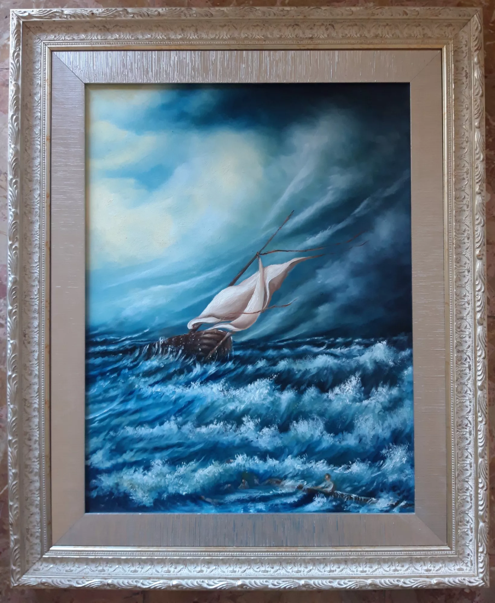 Dio-ci-salverà-Aly-Mohsen "Quadro di Ay Mohsen realizzato ad Olio con pennello. Il titolo è Dio ci salverà e rientra nella categoria Il mare. Opera originale di Aly Mohsen che raffigura un paesaggio marino in tempesta, caratterizzato da una forte tensione visiva e da una marcata dimensione simbolica. La scena è dominata da un mare agitato, costruito attraverso onde ampie e frante che occupano gran parte della composizione, mentre una barca solitaria, inclinata dalla forza del vento, avanza con difficoltà tra i flutti, diventando elemento centrale del racconto visivo. In primo piano, tra le onde, si distinguono figure umane appena percepibili, immerse nell’acqua in movimento. La loro presenza introduce una componente narrativa esplicita e rafforza il carattere drammatico dell’opera, trasformando il paesaggio in una scena di confronto diretto tra l’uomo e la potenza della natura. Il rapporto tra la piccola imbarcazione, i naufraghi e l’immensità del mare genera una composizione densa di significato e di tensione emotiva. Il cielo tempestoso è costruito con ampie campiture dinamiche, in cui i blu profondi e i grigi si stratificano creando un vortice di nuvole in movimento. Al centro della scena, una zona di luce più chiara si apre progressivamente tra le nubi, suggerendo un cambiamento atmosferico e introducendo un forte contrasto luministico. Questa apertura verso la luce guida lo sguardo e bilancia visivamente la violenza del mare sottostante. La tecnica pittorica evidenzia una piena maturità espressiva: l’uso della spatola e delle pennellate materiche conferisce alle onde una marcata tridimensionalità e un senso di moto continuo. Gli spessori cromatici e le variazioni tonali contribuiscono a rendere il mare vibrante e instabile, mentre la composizione complessiva mantiene un equilibrio strutturale rigoroso. L’opera è completata da una cornice elaborata di gusto classico, con un ampio passe-partout argentato dalla superficie finemente lavorata. La cornice dialoga con i toni freddi e profondi del dipinto, incorniciando la scena con eleganza e contribuendo all’unità visiva e alla presenza complessiva dell’opera."