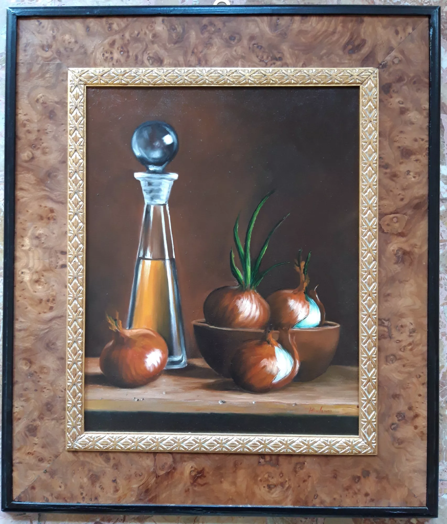 Oil-cruet-with-onions-Aly-Mohsen "Painting by Aly Mohsen, executed in Oil on canvas (brush). The title is Oil cruet with onions and it belongs to the category Still Life. Oil cruet with onions by Aly Mohsen is a figurative still life of restrained formal elegance, in which the artist addresses an everyday subject through a sober and carefully controlled pictorial language. The work belongs to the European still life tradition, reinterpreted with a contemporary sensitivity focused on light, material presence, and the spatial relationships between objects. The composition is essential and rigorously balanced: at the center of the scene stands a transparent glass oil cruet, filled with golden oil, which functions as the vertical focal point of the painting. Surrounding it, copper-toned onions are arranged on a wooden surface and gathered in a simple bowl, establishing a harmonious dialogue of rounded volumes, smooth surfaces, and natural textures. Delicate green shoots emerge subtly, introducing a sense of vitality against the dark, neutral background. The light is soft and precisely calibrated, enhancing the transparency of the glass, the chromatic density of the oil, and the glossy skins of the onions, resulting in a rendering that is both realistic and contemplative. The warm palette—dominated by browns, amber tones, and deep greens—contributes to an intimate, quiet atmosphere consistent with the tradition of figurative still life painting. Executed with the brush and marked by strong technical control, the painting reveals particular attention to material rendering and spatial clarity, confirming Aly Mohsen’s stylistic maturity in the treatment of simple, everyday subjects. The painting is completed by a high-quality frame featuring a wide outer band in burl wood, an inner rebate finely decorated with gold leaf, and a dark inner profile that contains the composition. This structure surrounds the work with balance and elegance, reinforcing its museum-style presentation."