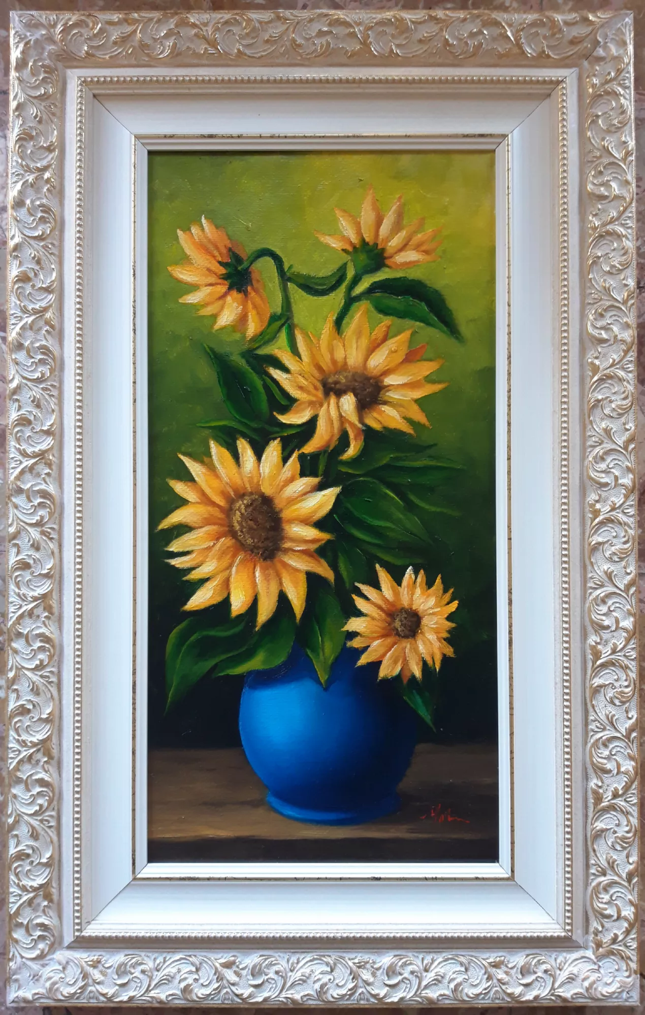 Vase-with-Sunflowers-Aly-Mohsen "Painting by Aly Mohsen, executed in Oil on canvas (brush). The title is Vase with Sunflowers and it belongs to the category Still Life. Vase with Sunflowers by Aly Mohsen is a floral still life distinguished by its clear compositional structure and by the intense chromatic relationship between subject and background. The work belongs to the tradition of figurative painting, approaching the floral theme through a sober and luminous pictorial language, with close attention to light and to the vitality of natural forms. The composition depicts a bouquet of sunflowers naturally arranged in a deep blue vase, placed on a horizontal surface. The vase acts as the central visual محور of the painting, creating a strong chromatic contrast with the warm yellows of the petals and the deep greens of the leaves. The flowers, shown at different angles and stages of bloom, introduce a dynamic rhythm that brings balance and movement to the composition. The green background, uniform and softly nuanced, envelops the scene without narrative distraction, enhancing the luminosity of the sunflowers and reinforcing the sense of spatial depth. The light, diffused and carefully controlled, models the petals and leaves with gradual transitions, emphasizing their material structure and volume. Executed with the brush, the painting demonstrates assured technique and careful attention to surface rendering and tonal modulation. The work reflects Aly Mohsen’s sustained interest in natural subjects as a field for formal and chromatic exploration, within the framework of still life painting interpreted through a contemporary sensibility. The painting is completed by a classically styled frame featuring a wide molding in light tones with raised ornamental motifs, which surrounds the work with balance and elegance, accentuates its vertical format, and reinforces its museum-style presentation."