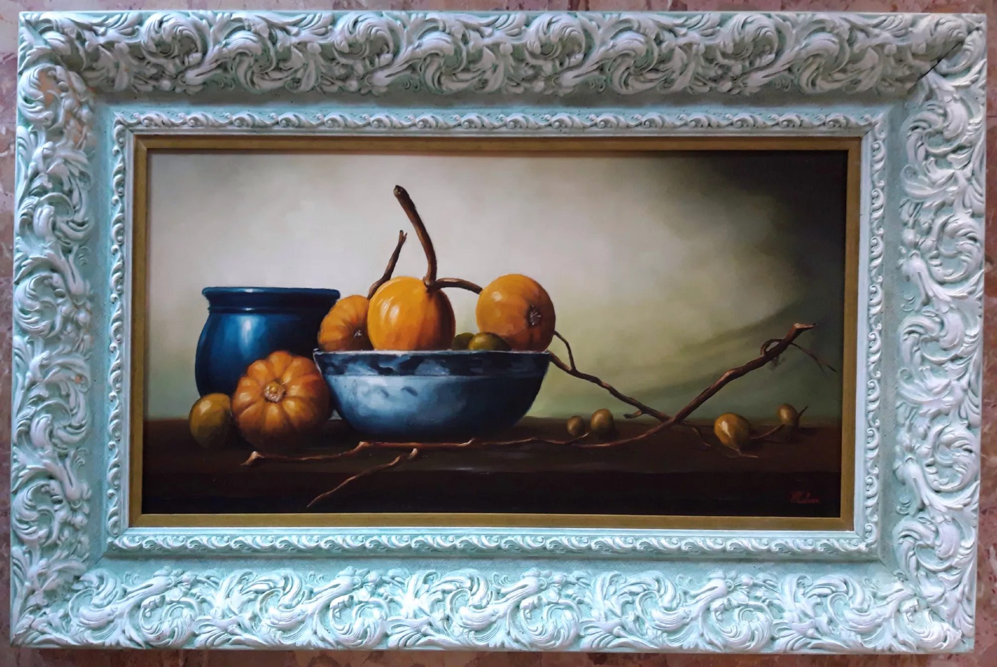 Still-Life-with-Pumpkins-Aly-Mohsen "Painting by Aly Mohsen, executed in Oil on canvas (brush). The title is Still Life with Pumpkins and it belongs to the category Still Life. The work is conceived as a still life within the context of figurative painting by Aly Mohsen, focused on a carefully arranged group of vegetal elements placed on a horizontal surface within a restrained and quiet pictorial space. The composition centers on a cluster of pumpkins in warm, golden tones, positioned inside and around a bowl and a ceramic vessel marked by deep blue hues, set against a neutral background. The fruits are accompanied by slender, sinuous branches and small vegetal details that extend across the pictorial surface, establishing a continuous and measured visual line. The arrangement of the objects reflects a balanced relationship between solids and voids, with controlled spacing and subtle overlaps. The light, soft and directional, shapes the volumes without sharp contrasts, emphasizing the distinct material qualities: the uneven skin of the pumpkins, the smooth glazed surface of the ceramic vessels, and the solidity of the dark supporting plane. The background, rendered through gradual tonal transitions, isolates the subject and enhances the sense of spatial depth. The execution reveals a restrained control of gesture, with paint application alternating between more compact areas and softer transitions, achieved through the use of the brush and a deliberate handling of pictorial matter. The frame, made of carved wood with a light finish and relief decoration, is completed by a gilded inner profile that defines the boundary of the image and reinforces its spatial perception, serving both a visual and structural framing function."