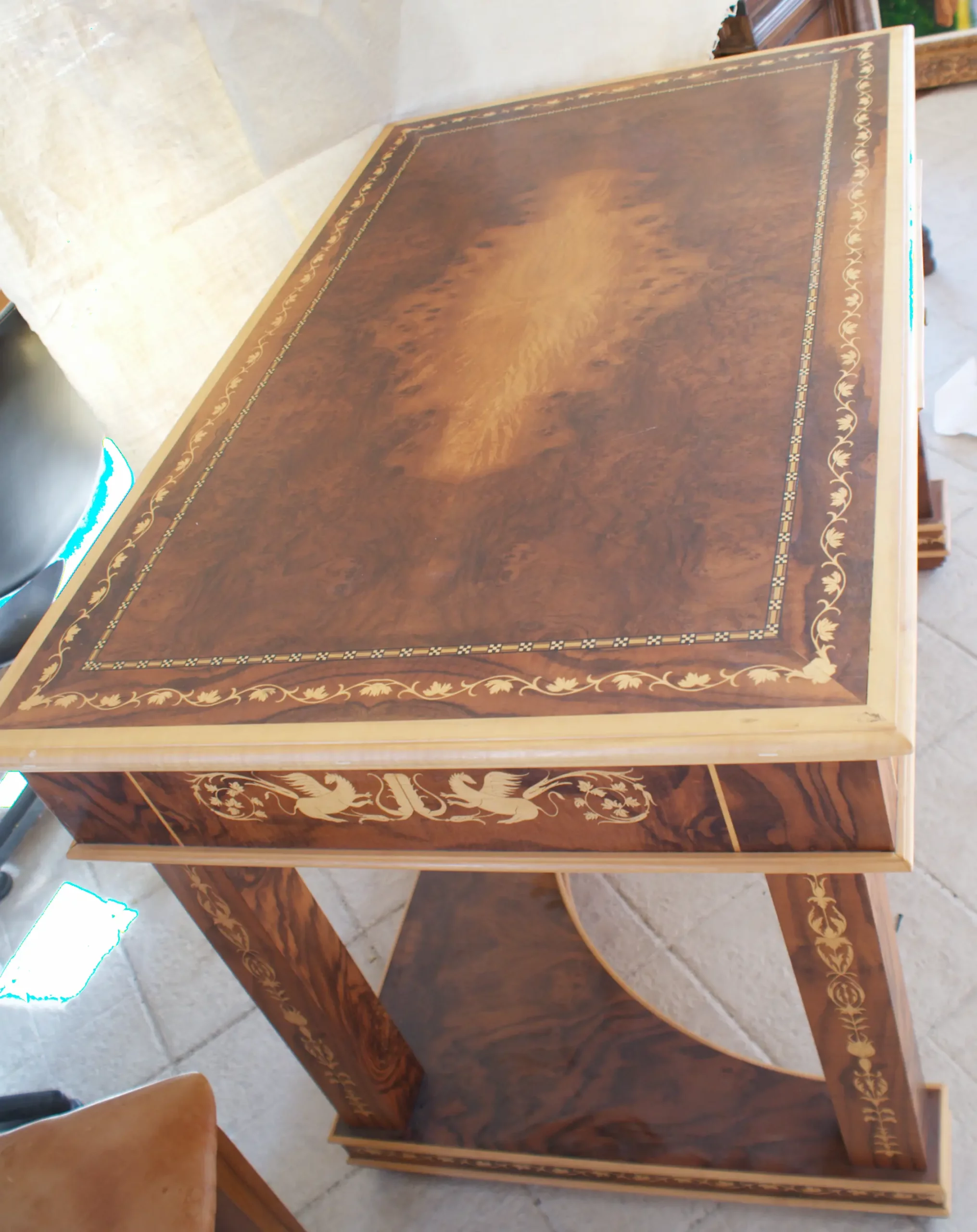 "Artwork by Aly Mohsen executed in the marquetry technique. The title is Scrivania delle chimere (Desk of the Chimeras – original Italian title) and it belongs to the category Monumental Cabinetmaking. The Scrivania delle Chimere is a monumental work of artistic cabinetmaking by Aly Mohsen, conceived as a unique, one-of-a-kind piece and executed in accordance with the traditions of high European fine woodworking. The work occupies a space between functional sculpture and symbolic artifact, integrating structure, decoration, and iconography into a unified artistic whole. The desktop is crafted from carefully selected walnut burl, distinguished by its complex, symmetrical grain pattern, enhanced through a refined finish that emphasizes both chromatic depth and material presence. The surface is framed by a polychrome marquetry border composed of white maple, ebony, and padauk, arranged in a continuous classical motif and executed with a high degree of technical precision. The supporting structure is made of solid mahogany, used for the legs, aprons, and primary architectural elements. Vertical surfaces are enriched with hand-cut white maple inlays featuring ornamental and figurative motifs that engage in a balanced dialogue with the overall structure. The curved lower base and the connecting elements between the legs contribute to both the visual stability and the structural coherence of the piece. A defining feature of the work is the presence of winged chimeras inlaid along the side panels. These figures carry an iconographic and symbolic significance, evoking themes of protection, strength, and transformation—recurring elements within Mohsen’s artistic research. Their inclusion is not merely decorative, but integral to the conceptual and narrative framework of the desk. The desk is conceived as a functional artwork, in which practical use is subordinate to an overarching artistic vision. The quality of the materials, the entirely hand-executed craftsmanship, and the unity between structure and decoration establish the piece as an autonomous artistic object, intended for representational settings where it can be experienced and interpreted as a work of art in its own right."