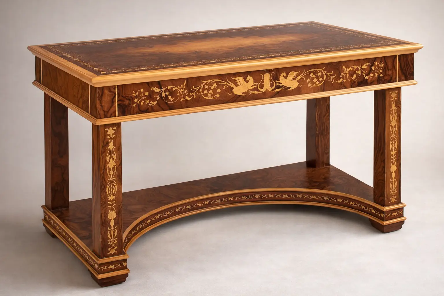 Scrivania-delle-chimere (Desk-of-the-Chimeras-original-Italian-title)-Aly-Mohsen "Artwork by Aly Mohsen executed in the marquetry technique. The title is Scrivania delle chimere (Desk of the Chimeras – original Italian title) and it belongs to the category Monumental Cabinetmaking. The Scrivania delle Chimere is a monumental work of artistic cabinetmaking by Aly Mohsen, conceived as a unique, one-of-a-kind piece and executed in accordance with the traditions of high European fine woodworking. The work occupies a space between functional sculpture and symbolic artifact, integrating structure, decoration, and iconography into a unified artistic whole. The desktop is crafted from carefully selected walnut burl, distinguished by its complex, symmetrical grain pattern, enhanced through a refined finish that emphasizes both chromatic depth and material presence. The surface is framed by a polychrome marquetry border composed of white maple, ebony, and padauk, arranged in a continuous classical motif and executed with a high degree of technical precision. The supporting structure is made of solid mahogany, used for the legs, aprons, and primary architectural elements. Vertical surfaces are enriched with hand-cut white maple inlays featuring ornamental and figurative motifs that engage in a balanced dialogue with the overall structure. The curved lower base and the connecting elements between the legs contribute to both the visual stability and the structural coherence of the piece. A defining feature of the work is the presence of winged chimeras inlaid along the side panels. These figures carry an iconographic and symbolic significance, evoking themes of protection, strength, and transformation—recurring elements within Mohsen’s artistic research. Their inclusion is not merely decorative, but integral to the conceptual and narrative framework of the desk. The desk is conceived as a functional artwork, in which practical use is subordinate to an overarching artistic vision. The quality of the materials, the entirely hand-executed craftsmanship, and the unity between structure and decoration establish the piece as an autonomous artistic object, intended for representational settings where it can be experienced and interpreted as a work of art in its own right."