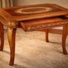 "Artwork by Aly Mohsen executed in the marquetry technique. The title is Scrivania degli angioletti (Desk of the Little Angels – original Italian title) and it belongs to the category Monumental Cabinetmaking. This unique work of artistic cabinetmaking belongs to the field of applied arts and reflects a synthesis of function and formal research within the production of Aly Mohsen. Conceived as an autonomous object, the desk is executed using traditional handcraft techniques of woodworking and marquetry, characterized by a restrained monumental presence. The structure is made of Italian walnut, selected for its structural stability and chromatic depth, combined with rosewood in warmer, reddish-brown tones used to define the main surfaces and volumes. The octagonal tabletop features a central white maple inlay, entirely hand-crafted, arranged in a classically inspired ornamental pattern and framed by a perimeter border that visually organizes the surface. The side panels and sculpted legs are further enriched with white maple inlays depicting allegorical figures and floral motifs developed in a rhythmic sequence. Ornamentation is integrated into the structure rather than applied to it, reinforcing the coherence between architectural form and decorative language. The careful combination of wood species creates a controlled chromatic balance, while the restrained finish is intended to enhance the natural grain of the materials and the precision of the marquetry, avoiding any surface emphasis. The desk is conceived as a functional work of art, suitable for representative interiors, exhibition spaces, or environments dedicated to the applied arts, and stands as a testimony to a cabinetmaking practice grounded in manual craftsmanship, unified design, and continuity with the European historical tradition."