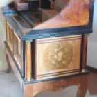 "Opera di Ay Mohsen realizzato con la tecnica dell'intarsio. Il titolo è Scrittoio della musica e rientra nella categoria Ebanisteria Monumentale. Questo straordinario Scrittoio della Musica, firmato Aly Mohsen, rappresenta uno dei vertici assoluti della sua produzione di ebanisteria artistica, in cui pittura, scultura e intarsio ligneo convivono in un unico oggetto funzionale e fortemente simbolico. L’opera si configura come un mobile scultoreo d’arte, concepito non come semplice arredo ma come opera autonoma, capace di dialogare con la tradizione delle arti applicate europee di alto livello. La struttura architettonica richiama con coerenza il linguaggio neoclassico e Impero, attraverso volumi pieni, una raffinata alzata centrale ad arco e colonne tornite coronate da capitelli in bronzo brunito, elementi che conferiscono solennità, ritmo e autorevolezza all’insieme. Il dialogo tra rigore formale e ricchezza decorativa costituisce uno dei tratti distintivi dell’opera. I pannelli frontali, laterali e posteriori sono impreziositi da intarsi figurativi a tema musicale e ornamentale: strumenti, spartiti, ghirlande e una raffinata composizione floreale in vaso, simboli universali di armonia, creatività e bellezza senza tempo. Particolarmente significativa è la decorazione del retro, concepita come un’opera completa e autonoma, scelta rara e consapevole che ribadisce la natura artistica totale dello scrittoio. Dal punto di vista materico, l’opera è realizzata attraverso una sapiente combinazione di radica di noce e radica di thuya, selezionate per la profondità cromatica e la vivacità delle venature. Acero, ebano e padouk sono impiegati per cornici, filettature e contrasti cromatici, mentre il pero definisce con eleganza la cornice dell’intarsio posteriore, introducendo una tonalità calda e luminosa che armonizza l’intera composizione. Il piano di scrittura estraibile, scandito da campiture scure e filetti chiari, incornicia un riquadro centrale intarsiato che funge da fulcro visivo e funzionale. I numerosi cassetti, perfettamente integrati nella struttura, testimoniano l’elevatissimo livello tecnico dell’artista e la sua capacità di coniugare complessità costruttiva ed equilibrio formale. Interamente realizzato a mano, senza alcun ricorso a processi industriali, lo Scrittoio della Musica si impone come testimonianza dell’alta ebanisteria artistica europea contemporanea, in cui si rinnova consapevolmente la grande tradizione delle arti applicate storiche. L’opera si colloca idealmente tra arte applicata, scultura funzionale e patrimonio culturale, risultando idonea a contesti museali, collezioni storiche e ambienti di rappresentanza di alto profilo culturale."