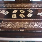 "Opera di Ay Mohsen realizzata con la tecnica dell'intarsio. Il titolo è Scrivania del poker e le tre carte e rientra nella categoria Ebanisteria Monumentale. Questa imponente scrivania monumentale rappresenta una delle opere più concettualmente audaci, raffinate e politicamente consapevoli di Aly Mohsen. In essa, la grande tradizione dell’ebanisteria artistica europea viene trasformata in strumento narrativo, simbolico e apertamente critico, dando vita a un’opera che trascende la funzione d’arredo per assumere la forma di messaggio cifrato e denuncia silenziosa. Non si tratta di un semplice mobile funzionale, ma di una vera opera d’arte tridimensionale, concepita per dialogare con il potere, smascherarne i meccanismi e mettere in crisi le apparenze di legalità e correttezza. Struttura e materiali La scrivania è caratterizzata da una struttura solida ma visivamente equilibrata, con gambe sagomate e slanciate che alleggeriscono la massa monumentale del piano. L’opera è realizzata con una selezione estremamente ricercata di legni pregiati, scelti non solo per le qualità estetiche ma anche per il loro valore simbolico: Noce nazionale e noce canaletto per la struttura principale e le superfici estese, apprezzati per la profondità cromatica e la stabilità nel tempo Radica di noce per i pannelli frontali, impiegata per ottenere superfici vibranti e irregolari, metafora visiva dell’instabilità e dell’inganno Ebano ed essenze scure tinte per i fondi degli intarsi, funzionali a rafforzare il contrasto grafico Acero e acero sicomoro per gli intarsi chiari Legni naturalmente colorati (padouk, acero, mogano, rovere, radica di noce, ebano verde ed ebano nero) per la resa cromatica delle figure e delle carte da gioco Il bordo chiaro e continuo del piano richiama deliberatamente la tradizione dei grandi tavoli da gioco, incorniciando la scena e convogliando lo sguardo verso il centro narrativo. Il piano: il gioco come metafora del potere Il piano superiore costituisce il cuore concettuale dell’opera. All’interno di una ricchissima cornice ornamentale intarsiata si sviluppa una sequenza di carte da poker disposte con apparente casualità. A una prima lettura, l’immagine suggerisce una partita in corso. A uno sguardo più attento, emerge però un elemento perturbante: le combinazioni non conducono a nessun esito logico. Le mani sono incoerenti, impossibili, volutamente manipolate. Questa ambiguità è intenzionale. Il poker, simbolo di strategia, calcolo e rischio controllato, viene qui messo in relazione con il gioco delle tre carte, archetipo universale della truffa. Il messaggio è sottile ma inequivocabile: chi crede di giocare secondo regole condivise è già vittima di un inganno. Il fronte: musica, scena e finzione Il fronte della scrivania è dominato da un grande medaglione circolare intarsiato, nel quale compaiono strumenti musicali, spartiti, carte da gioco e motivi floreali. La musica diventa metafora della messa in scena del potere: un concerto apparentemente armonioso che nasconde dissonanze profonde, una composizione elegante pensata per distrarre, sedurre e confondere. Le fasce laterali, ornate con ghirlande classiche e figure stilizzate, richiamano consapevolmente il linguaggio neoclassico e ottocentesco, rafforzando l’idea di ordine, autorità e rispettabilità — un ordine solo apparente. Storia dell’opera e valore simbolico La scrivania fu commissionata da un importante uomo d’affari egiziano, figura di enorme peso economico internazionale. Aly Mohsen, che conosceva il committente fin dall’infanzia, aveva fondati motivi per ritenere che questi operasse in realtà come prestanome per i servizi segreti statunitensi, coinvolto in operazioni economiche e finanziarie condotte in Italia. Al momento della consegna, Mohsen si recò personalmente nell’ufficio del committente e ne spiegò il significato con parole che rivelano la natura profonda dell’opera: «Questa scrivania serve per avvisare chi si siede di fronte a te: lui pensa di giocare a poker, mentre tu stai giocando al gioco delle tre carte.» Un’accusa di straordinaria eleganza, affidata non al linguaggio diretto ma alla forza simbolica dell’intarsio. Il committente, comprendendo perfettamente il messaggio, si sentì apertamente insultato e rifiutò l’opera. La scrivania rimase così nelle mani dell’artista. Rimasta nella disponibilità dell’artista, la Scrivania del Poker e del Gioco delle Tre Carte possiede oggi un valore ulteriormente accresciuto. È un pezzo unico, documentato, con una storia forte, verificabile e di straordinaria attrattiva per il collezionismo internazionale. L’opera unisce: alta ebanisteria artistica contenuto politico e narrativo qualità museale dimensioni monumentali provenienza diretta dall’artista Per collezionisti evoluti e investitori in mobili d’arte, arti decorative e opere concettuali, questa scrivania rappresenta un caso raro di arredo che diventa linguaggio critico, capace di dialogare con design, scultura e arte contemporanea, e destinato a una progressiva valorizzazione culturale e patrimoniale. In Aly Mohsen, il legno non è mai solo materia: diventa linguaggio, accusa, ironia e memoria. E questa scrivania ne è una delle dichiarazioni più lucide e implacabili."