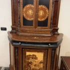 "Opera di Ay Mohsen realizzata con la tecnica dell'intarsio. Il titolo è Credenza del ponte vecchio e rientra nella categoria Ebanisteria Monumentale. La Credenza del Ponte Vecchio di Ivrea è un mobile artistico intarsiato e scolpito concepito come opera unica di alta ebanisteria, nel quale confluiscono tradizione figurativa, lavorazione lignea d’autore e riferimento paesaggistico di carattere identitario. L’opera si configura come un manufatto tridimensionale complesso, in cui struttura architettonica, decorazione plastica e apparato intarsiato concorrono alla definizione di un insieme unitario. La struttura è realizzata in mogano massello e radica di noce, utilizzati per le parti portanti e per le superfici principali, con un’alternanza di campiture compatte e venature irregolari che articolano il volume complessivo. Le fiancate presentano elementi scultorei in noce, interamente scolpiti a mano, raffiguranti figure stilizzate integrate verticalmente nei montanti, che accentuano il carattere plastico e monumentale del mobile e richiamano la tradizione europea dell’intaglio figurativo. Il bordo superiore e il cassetto sono realizzati in ebano, introducendo un contrasto cromatico netto con le essenze più calde della struttura. Le ante superiori presentano fondi scuri in ebano sui quali si sviluppano intarsi floreali policromi, eseguiti in acero, noce, ebano verde e ciliegio, organizzati in composizioni simmetriche che definiscono un equilibrio tra decorazione e ordine geometrico. Elemento centrale dell’opera è il grande pannello intarsiato posto nella parte inferiore del mobile, raffigurante una veduta del Ponte Vecchio di Ivrea. La scena paesaggistica è costruita mediante una complessa combinazione di essenze lignee, tra cui acero, radica di noce, noce chiaro e scuro ed ebano verde, utilizzate per ottenere variazioni tonali, profondità spaziale e articolazione luministica. Il riquadro è delimitato da una cornice in legno di rose che ne definisce il perimetro e ne isola visivamente l’immagine dal resto della struttura. L’interno del mobile è completamente rifinito e decorato, con pannelli intarsiati a motivi floreali e superfici arricchite da disegni geometrici a raggiera e campiture angolari ottenute mediante l’accostamento di diverse essenze lignee. Le decorazioni interne sono concepite come parte integrante dell’opera e risultano pienamente visibili grazie alla presenza di un ripiano in vetro, che consente la lettura continua degli intarsi anche a mobile aperto. La credenza si conclude con una cornice strutturale continua che accompagna i profili superiori e inferiori del mobile, realizzata in essenza scura con finitura uniforme, avente la funzione di contenere visivamente il volume complessivo e di raccordare le diverse superfici lignee in un insieme coerente e unitario."