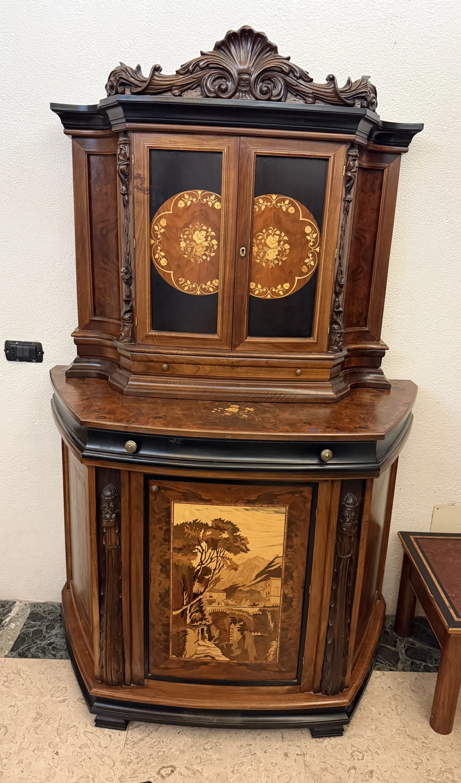 "Opera di Ay Mohsen realizzata con la tecnica dell'intarsio. Il titolo è Credenza del ponte vecchio e rientra nella categoria Ebanisteria Monumentale. La Credenza del Ponte Vecchio di Ivrea è un mobile artistico intarsiato e scolpito concepito come opera unica di alta ebanisteria, nel quale confluiscono tradizione figurativa, lavorazione lignea d’autore e riferimento paesaggistico di carattere identitario. L’opera si configura come un manufatto tridimensionale complesso, in cui struttura architettonica, decorazione plastica e apparato intarsiato concorrono alla definizione di un insieme unitario. La struttura è realizzata in mogano massello e radica di noce, utilizzati per le parti portanti e per le superfici principali, con un’alternanza di campiture compatte e venature irregolari che articolano il volume complessivo. Le fiancate presentano elementi scultorei in noce, interamente scolpiti a mano, raffiguranti figure stilizzate integrate verticalmente nei montanti, che accentuano il carattere plastico e monumentale del mobile e richiamano la tradizione europea dell’intaglio figurativo. Il bordo superiore e il cassetto sono realizzati in ebano, introducendo un contrasto cromatico netto con le essenze più calde della struttura. Le ante superiori presentano fondi scuri in ebano sui quali si sviluppano intarsi floreali policromi, eseguiti in acero, noce, ebano verde e ciliegio, organizzati in composizioni simmetriche che definiscono un equilibrio tra decorazione e ordine geometrico. Elemento centrale dell’opera è il grande pannello intarsiato posto nella parte inferiore del mobile, raffigurante una veduta del Ponte Vecchio di Ivrea. La scena paesaggistica è costruita mediante una complessa combinazione di essenze lignee, tra cui acero, radica di noce, noce chiaro e scuro ed ebano verde, utilizzate per ottenere variazioni tonali, profondità spaziale e articolazione luministica. Il riquadro è delimitato da una cornice in legno di rose che ne definisce il perimetro e ne isola visivamente l’immagine dal resto della struttura. L’interno del mobile è completamente rifinito e decorato, con pannelli intarsiati a motivi floreali e superfici arricchite da disegni geometrici a raggiera e campiture angolari ottenute mediante l’accostamento di diverse essenze lignee. Le decorazioni interne sono concepite come parte integrante dell’opera e risultano pienamente visibili grazie alla presenza di un ripiano in vetro, che consente la lettura continua degli intarsi anche a mobile aperto. La credenza si conclude con una cornice strutturale continua che accompagna i profili superiori e inferiori del mobile, realizzata in essenza scura con finitura uniforme, avente la funzione di contenere visivamente il volume complessivo e di raccordare le diverse superfici lignee in un insieme coerente e unitario."