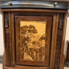 "Opera di Ay Mohsen realizzata con la tecnica dell'intarsio. Il titolo è Credenza del ponte vecchio e rientra nella categoria Ebanisteria Monumentale. La Credenza del Ponte Vecchio di Ivrea è un mobile artistico intarsiato e scolpito concepito come opera unica di alta ebanisteria, nel quale confluiscono tradizione figurativa, lavorazione lignea d’autore e riferimento paesaggistico di carattere identitario. L’opera si configura come un manufatto tridimensionale complesso, in cui struttura architettonica, decorazione plastica e apparato intarsiato concorrono alla definizione di un insieme unitario. La struttura è realizzata in mogano massello e radica di noce, utilizzati per le parti portanti e per le superfici principali, con un’alternanza di campiture compatte e venature irregolari che articolano il volume complessivo. Le fiancate presentano elementi scultorei in noce, interamente scolpiti a mano, raffiguranti figure stilizzate integrate verticalmente nei montanti, che accentuano il carattere plastico e monumentale del mobile e richiamano la tradizione europea dell’intaglio figurativo. Il bordo superiore e il cassetto sono realizzati in ebano, introducendo un contrasto cromatico netto con le essenze più calde della struttura. Le ante superiori presentano fondi scuri in ebano sui quali si sviluppano intarsi floreali policromi, eseguiti in acero, noce, ebano verde e ciliegio, organizzati in composizioni simmetriche che definiscono un equilibrio tra decorazione e ordine geometrico. Elemento centrale dell’opera è il grande pannello intarsiato posto nella parte inferiore del mobile, raffigurante una veduta del Ponte Vecchio di Ivrea. La scena paesaggistica è costruita mediante una complessa combinazione di essenze lignee, tra cui acero, radica di noce, noce chiaro e scuro ed ebano verde, utilizzate per ottenere variazioni tonali, profondità spaziale e articolazione luministica. Il riquadro è delimitato da una cornice in legno di rose che ne definisce il perimetro e ne isola visivamente l’immagine dal resto della struttura. L’interno del mobile è completamente rifinito e decorato, con pannelli intarsiati a motivi floreali e superfici arricchite da disegni geometrici a raggiera e campiture angolari ottenute mediante l’accostamento di diverse essenze lignee. Le decorazioni interne sono concepite come parte integrante dell’opera e risultano pienamente visibili grazie alla presenza di un ripiano in vetro, che consente la lettura continua degli intarsi anche a mobile aperto. La credenza si conclude con una cornice strutturale continua che accompagna i profili superiori e inferiori del mobile, realizzata in essenza scura con finitura uniforme, avente la funzione di contenere visivamente il volume complessivo e di raccordare le diverse superfici lignee in un insieme coerente e unitario."