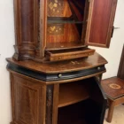 "Opera di Ay Mohsen realizzata con la tecnica dell'intarsio. Il titolo è Credenza del ponte vecchio e rientra nella categoria Ebanisteria Monumentale. La Credenza del Ponte Vecchio di Ivrea è un mobile artistico intarsiato e scolpito concepito come opera unica di alta ebanisteria, nel quale confluiscono tradizione figurativa, lavorazione lignea d’autore e riferimento paesaggistico di carattere identitario. L’opera si configura come un manufatto tridimensionale complesso, in cui struttura architettonica, decorazione plastica e apparato intarsiato concorrono alla definizione di un insieme unitario. La struttura è realizzata in mogano massello e radica di noce, utilizzati per le parti portanti e per le superfici principali, con un’alternanza di campiture compatte e venature irregolari che articolano il volume complessivo. Le fiancate presentano elementi scultorei in noce, interamente scolpiti a mano, raffiguranti figure stilizzate integrate verticalmente nei montanti, che accentuano il carattere plastico e monumentale del mobile e richiamano la tradizione europea dell’intaglio figurativo. Il bordo superiore e il cassetto sono realizzati in ebano, introducendo un contrasto cromatico netto con le essenze più calde della struttura. Le ante superiori presentano fondi scuri in ebano sui quali si sviluppano intarsi floreali policromi, eseguiti in acero, noce, ebano verde e ciliegio, organizzati in composizioni simmetriche che definiscono un equilibrio tra decorazione e ordine geometrico. Elemento centrale dell’opera è il grande pannello intarsiato posto nella parte inferiore del mobile, raffigurante una veduta del Ponte Vecchio di Ivrea. La scena paesaggistica è costruita mediante una complessa combinazione di essenze lignee, tra cui acero, radica di noce, noce chiaro e scuro ed ebano verde, utilizzate per ottenere variazioni tonali, profondità spaziale e articolazione luministica. Il riquadro è delimitato da una cornice in legno di rose che ne definisce il perimetro e ne isola visivamente l’immagine dal resto della struttura. L’interno del mobile è completamente rifinito e decorato, con pannelli intarsiati a motivi floreali e superfici arricchite da disegni geometrici a raggiera e campiture angolari ottenute mediante l’accostamento di diverse essenze lignee. Le decorazioni interne sono concepite come parte integrante dell’opera e risultano pienamente visibili grazie alla presenza di un ripiano in vetro, che consente la lettura continua degli intarsi anche a mobile aperto. La credenza si conclude con una cornice strutturale continua che accompagna i profili superiori e inferiori del mobile, realizzata in essenza scura con finitura uniforme, avente la funzione di contenere visivamente il volume complessivo e di raccordare le diverse superfici lignee in un insieme coerente e unitario."