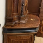 "Opera di Ay Mohsen realizzata con la tecnica dell'intarsio. Il titolo è Credenza del ponte vecchio e rientra nella categoria Ebanisteria Monumentale. La Credenza del Ponte Vecchio di Ivrea è un mobile artistico intarsiato e scolpito concepito come opera unica di alta ebanisteria, nel quale confluiscono tradizione figurativa, lavorazione lignea d’autore e riferimento paesaggistico di carattere identitario. L’opera si configura come un manufatto tridimensionale complesso, in cui struttura architettonica, decorazione plastica e apparato intarsiato concorrono alla definizione di un insieme unitario. La struttura è realizzata in mogano massello e radica di noce, utilizzati per le parti portanti e per le superfici principali, con un’alternanza di campiture compatte e venature irregolari che articolano il volume complessivo. Le fiancate presentano elementi scultorei in noce, interamente scolpiti a mano, raffiguranti figure stilizzate integrate verticalmente nei montanti, che accentuano il carattere plastico e monumentale del mobile e richiamano la tradizione europea dell’intaglio figurativo. Il bordo superiore e il cassetto sono realizzati in ebano, introducendo un contrasto cromatico netto con le essenze più calde della struttura. Le ante superiori presentano fondi scuri in ebano sui quali si sviluppano intarsi floreali policromi, eseguiti in acero, noce, ebano verde e ciliegio, organizzati in composizioni simmetriche che definiscono un equilibrio tra decorazione e ordine geometrico. Elemento centrale dell’opera è il grande pannello intarsiato posto nella parte inferiore del mobile, raffigurante una veduta del Ponte Vecchio di Ivrea. La scena paesaggistica è costruita mediante una complessa combinazione di essenze lignee, tra cui acero, radica di noce, noce chiaro e scuro ed ebano verde, utilizzate per ottenere variazioni tonali, profondità spaziale e articolazione luministica. Il riquadro è delimitato da una cornice in legno di rose che ne definisce il perimetro e ne isola visivamente l’immagine dal resto della struttura. L’interno del mobile è completamente rifinito e decorato, con pannelli intarsiati a motivi floreali e superfici arricchite da disegni geometrici a raggiera e campiture angolari ottenute mediante l’accostamento di diverse essenze lignee. Le decorazioni interne sono concepite come parte integrante dell’opera e risultano pienamente visibili grazie alla presenza di un ripiano in vetro, che consente la lettura continua degli intarsi anche a mobile aperto. La credenza si conclude con una cornice strutturale continua che accompagna i profili superiori e inferiori del mobile, realizzata in essenza scura con finitura uniforme, avente la funzione di contenere visivamente il volume complessivo e di raccordare le diverse superfici lignee in un insieme coerente e unitario."