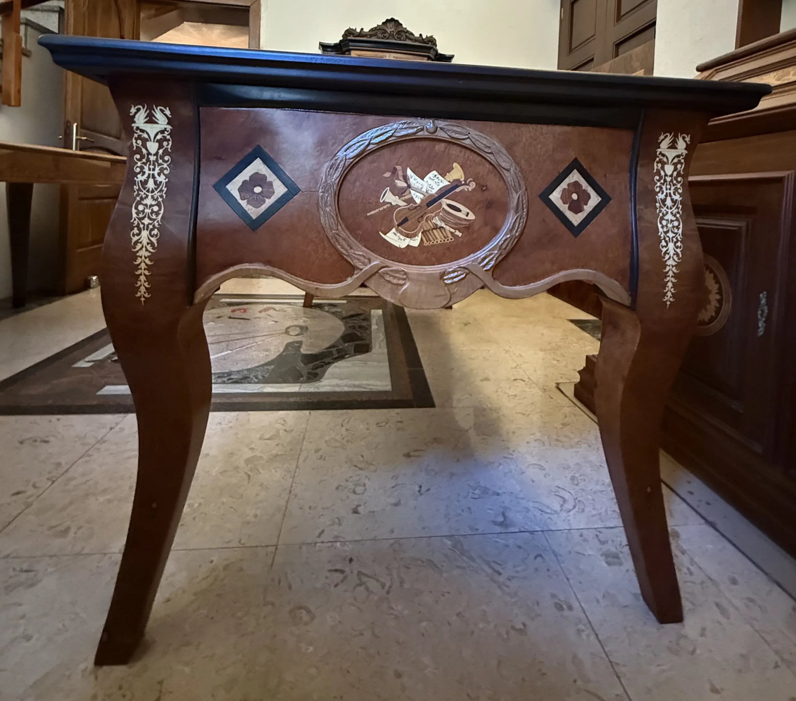 "Artwork by Aly Mohsen executed in the marquetry technique. The title is Scrivania dei fiori neri (Desk of the Black Flowers – original Italian title) and it belongs to the category Monumental Cabinetmaking. An artistic cabinetmaking desk conceived as a unique work in which functional use is integrated with an articulated decorative system of figurative character. The piece is situated within the field of contemporary authorial cabinetmaking, employing a formal language consistent with the historical tradition of wooden marquetry and hand-crafted woodworking. The tabletop is made of rosewood, with a central section in walnut burl arranged in mirrored grain along a longitudinal axis that organizes the entire composition. This central field is enclosed by a frame in walnut and white maple, within which geometric inlays in ebony, maple, and walnut are inserted. These elements are distributed in a regular manner and serve to define the visual space. The surface displays a chromatic range dominated by warm, brown tonalities, articulated through contrasts between light and dark woods. Dark-toned floral inlays are present on the tabletop, executed in ebony, maple, and padauk and arranged symmetrically along the perimeter. These elements introduce a marked chromatic contrast and contribute to the rhythmic articulation of the composition. The perimeter edge of the tabletop, executed in ebony, clearly defines the profile of the desk and reinforces its architectural reading. The legs, made of burl wood and shaped with a tapered profile, are decorated with linear white maple inlays that visually lighten their volume while maintaining formal continuity with the tabletop and side panels. The short sides feature diamond-shaped inlays with stylized floral motifs, while at the center of one of the long sides a figurative inlay depicting musical instruments is placed, composed through the juxtaposition of ebony, maple, and walnut. This inlay is enclosed by a hand-carved pearwood frame that separates the image from the structural field of the furniture. The opposite side features a figurative inlay depicting a female figure protecting a child, realized through layered woods of varying tonalities, with chiaroscuro effects achieved exclusively through chromatic variation of the materials. The composition is frontal and focused on the figures, which are set within a carved pearwood frame serving both to contain the image and to define the visual space of the figurative field. The structure includes five drawers aligned along the long side, integrated into the lower band without formal interruption, contributing to the horizontal articulation of the overall form. Light interacts with the polished surfaces and the different wood species, enhancing the depth of the grain and the legibility of the inlays, confirming the desk as a work in which cabinetmaking, figurative marquetry, and compositional organization converge into a single coherent object."