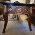 "Opera di Ay Mohsen realizzata con la tecnica dell'Intarsio. Il titolo è Scrivania dei fiori neri e rientra nella categoria Ebanisteria Monumentale. Scrivania di ebanisteria artistica, concepita come opera unica in cui la funzione d’uso è integrata a un articolato impianto decorativo di carattere figurativo. L’opera si colloca nell’ambito dell’ebanisteria d’autore contemporanea, con un linguaggio formale coerente con la tradizione storica dell’intarsio ligneo e della lavorazione manuale del legno. Il piano è realizzato in legno di rose, con una sezione centrale in radica di noce disposta a venatura speculare, secondo un asse longitudinale che organizza l’intera composizione. La campitura centrale è delimitata da una cornice in noce ed acero bianco, all’interno della quale si inseriscono intarsi geometrici in ebano, acero e noce, distribuiti in modo regolare e funzionali alla definizione dello spazio visivo. La superficie presenta una gamma cromatica dominata da toni caldi e bruni, modulati attraverso contrasti tra essenze chiare e scure. Sul piano sono presenti intarsi floreali di colore scuro, realizzati in ebano, acero e padouk, disposti simmetricamente lungo il perimetro. Tali elementi introducono un marcato contrasto cromatico e contribuiscono alla scansione ritmica della composizione. Il bordo perimetrale del piano, eseguito in ebano, definisce con chiarezza il profilo della scrivania e ne rafforza la lettura architettonica. Le gambe, realizzate in radica e sagomate con andamento rastremato, sono decorate da intarsi lineari in acero bianco che ne alleggeriscono visivamente il volume, mantenendo una continuità formale con il piano e con le fasce laterali. I lati corti presentano intarsi a rombo con motivi floreali stilizzati, mentre al centro di uno dei lati lunghi è collocato un intarsio figurativo raffigurante strumenti musicali, composto mediante l’accostamento di ebano, acero e noce. L’intarsio è delimitato da una cornice scolpita a mano in legno di pero, che separa l’immagine dal campo strutturale del mobile. Il lato opposto ospita un intarsio figurativo raffigurante una figura femminile che protegge un bambino, realizzato attraverso sovrapposizioni di essenze lignee di diversa tonalità, con una resa chiaroscurale affidata esclusivamente alla variazione cromatica dei materiali. La composizione è frontale e concentrata sulle figure, inserite entro una cornice scolpita in legno di pero, con funzione di contenimento dell’immagine e di definizione visiva dello spazio figurativo. La struttura include cinque cassetti allineati sul lato lungo, integrati nella fascia inferiore senza interruzioni formali, contribuendo alla scansione orizzontale dell’insieme. La luce interagisce con le superfici lucidate e con le diverse essenze lignee accentuando la profondità delle venature e la leggibilità degli intarsi, confermando la scrivania come opera in cui ebanisteria, intarsio figurativo e organizzazione compositiva convergono in un unico manufatto coerente."