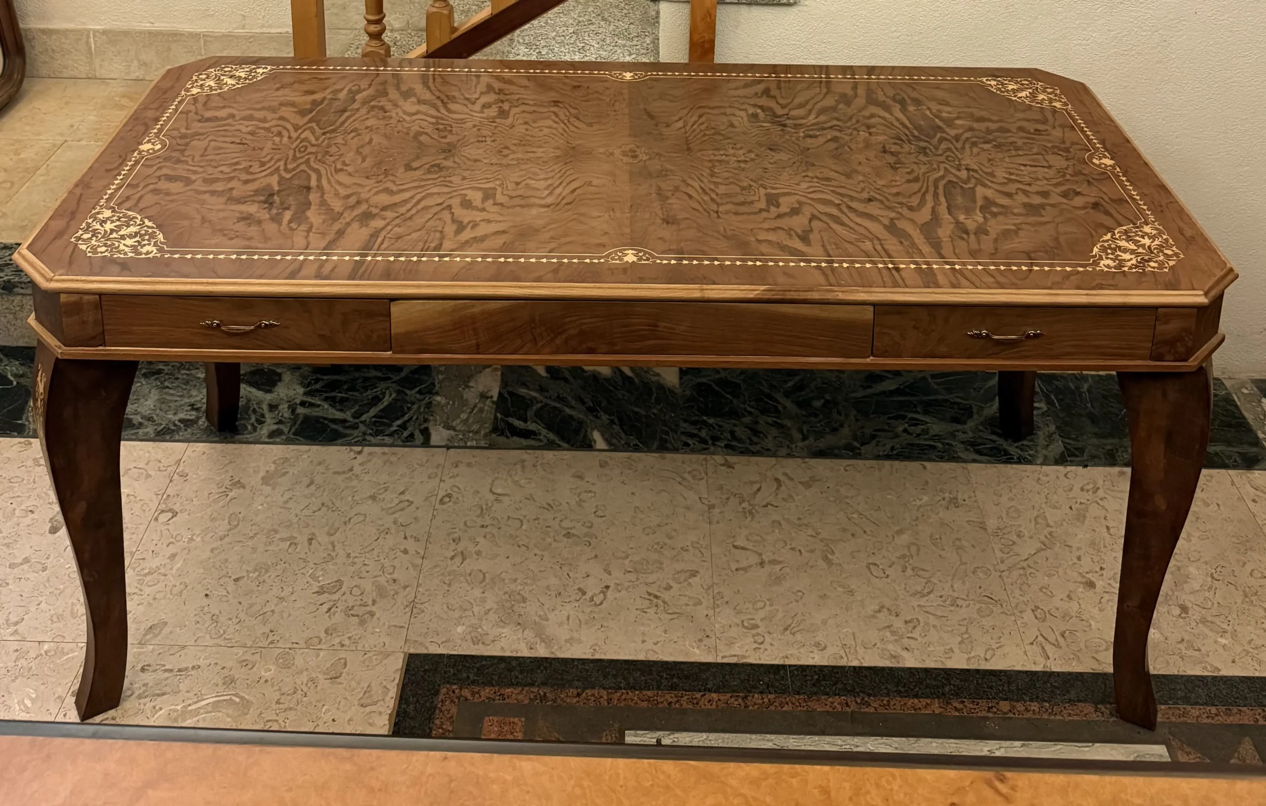 "Artwork by Aly Mohsen executed in the marquetry technique. The title is Neoclassical desk and it belongs to the category Monumental Cabinetmaking. This neoclassical inlaid desk by Aly Mohsen is conceived as a work of high artistic cabinetmaking, understood as a unified object in which function and formal construction are integrated within a decorative and architectural structure. The piece is rooted in the tradition of European classical furniture, reinterpreted through a controlled approach to materials and compositional order. The tabletop is crafted from selected mahogany feather veneer, distinguished by a symmetrical and continuous grain that defines a compact and visually coherent surface. The central field is framed by an articulated system of light maple inlays arranged in a neoclassical perimeter design, featuring floral and phytomorphic motifs positioned at the corners and along the midpoints of the sides. The relationship between the inlaid decoration and the natural figuring of the mahogany is structured through rhythmic continuity and visual balance. The edges of the tabletop are finished in light walnut, creating a gradual chromatic transition between surface and structure. The main body of the desk and the drawers are executed in dark walnut, providing visual solidity and tonal consistency while reinforcing the architectural reading of the piece. The detachable legs are constructed with a laminated mahogany core and subsequently veneered in oak burl. Each leg is decorated with vertical light maple inlays, conceived as continuous ornamental elements that emphasize vertical development and reinforce the sculptural character of the structure. The perimeter edge of the tabletop functions as a framing element for the composition, providing visual containment and spatial definition while delimiting the inlaid surface and contributing to the overall formal stability and perceptual coherence of the desk."
