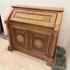 "Opera di Ay Mohsen realizzata con la tecnica dell'intarsio. Il titolo è Scrittoio delle ghirlande e rientra nella categoria Ebanisteria Monumentale. Lo Scrittoio delle Ghirlande di Aly Mohsen si configura come un mobile artistico intarsiato e scolpito, concepito come opera unica di alta ebanisteria, in cui funzione e articolazione decorativa sono integrate in una struttura di impianto architettonico. L’opera si colloca nella tradizione del mobile classico monumentale, reinterpretata attraverso una progettazione rigorosa e un controllo accurato dei materiali. La struttura portante è realizzata in noce calabrese della Sila, essenza selezionata per la compattezza, la stabilità strutturale e la profondità cromatica. I fianchi sagomati e le colonne frontali in radica di thuya introducono un ritmo plastico verticale, ulteriormente definito dalla presenza di elementi decorativi in bronzo che sottolineano i punti di raccordo e rafforzano la lettura architettonica del mobile. Elemento centrale dell’opera è il sistema di intarsi a ghirlanda, sviluppato secondo un disegno continuo e simmetrico. I profili degli intarsi sono eseguiti in acero, ebano e legno di rose, mentre i motivi floreali interni utilizzano una combinazione calibrata di acero, ebano verde, noce chiaro e noce scuro. La variazione cromatica tra le diverse essenze è costruita per gradazioni tonali e per equilibrio materico, in dialogo costante con il fondo ligneo. L’anta ribaltabile, incernierata sul fronte, rivela un interno organizzato con vani e cassetti in rovere, materiale scelto per la sua resistenza e per il contrasto visivo con le essenze esterne. Anche nella parte interna la lavorazione mantiene coerenza formale e qualità esecutiva, confermando l’unitarietà progettuale dell’opera. Il piano superiore e i bordi perimetrali sono trattati come una cornice strutturale e visiva, con funzione di contenimento dell’apparato decorativo e di definizione spaziale dell’insieme, contribuendo alla stabilità percettiva e alla chiarezza compositiva dello scrittoio."