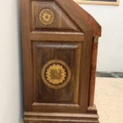 "Opera di Ay Mohsen realizzata con la tecnica dell'intarsio. Il titolo è Scrittoio delle ghirlande e rientra nella categoria Ebanisteria Monumentale. Lo Scrittoio delle Ghirlande di Aly Mohsen si configura come un mobile artistico intarsiato e scolpito, concepito come opera unica di alta ebanisteria, in cui funzione e articolazione decorativa sono integrate in una struttura di impianto architettonico. L’opera si colloca nella tradizione del mobile classico monumentale, reinterpretata attraverso una progettazione rigorosa e un controllo accurato dei materiali. La struttura portante è realizzata in noce calabrese della Sila, essenza selezionata per la compattezza, la stabilità strutturale e la profondità cromatica. I fianchi sagomati e le colonne frontali in radica di thuya introducono un ritmo plastico verticale, ulteriormente definito dalla presenza di elementi decorativi in bronzo che sottolineano i punti di raccordo e rafforzano la lettura architettonica del mobile. Elemento centrale dell’opera è il sistema di intarsi a ghirlanda, sviluppato secondo un disegno continuo e simmetrico. I profili degli intarsi sono eseguiti in acero, ebano e legno di rose, mentre i motivi floreali interni utilizzano una combinazione calibrata di acero, ebano verde, noce chiaro e noce scuro. La variazione cromatica tra le diverse essenze è costruita per gradazioni tonali e per equilibrio materico, in dialogo costante con il fondo ligneo. L’anta ribaltabile, incernierata sul fronte, rivela un interno organizzato con vani e cassetti in rovere, materiale scelto per la sua resistenza e per il contrasto visivo con le essenze esterne. Anche nella parte interna la lavorazione mantiene coerenza formale e qualità esecutiva, confermando l’unitarietà progettuale dell’opera. Il piano superiore e i bordi perimetrali sono trattati come una cornice strutturale e visiva, con funzione di contenimento dell’apparato decorativo e di definizione spaziale dell’insieme, contribuendo alla stabilità percettiva e alla chiarezza compositiva dello scrittoio."