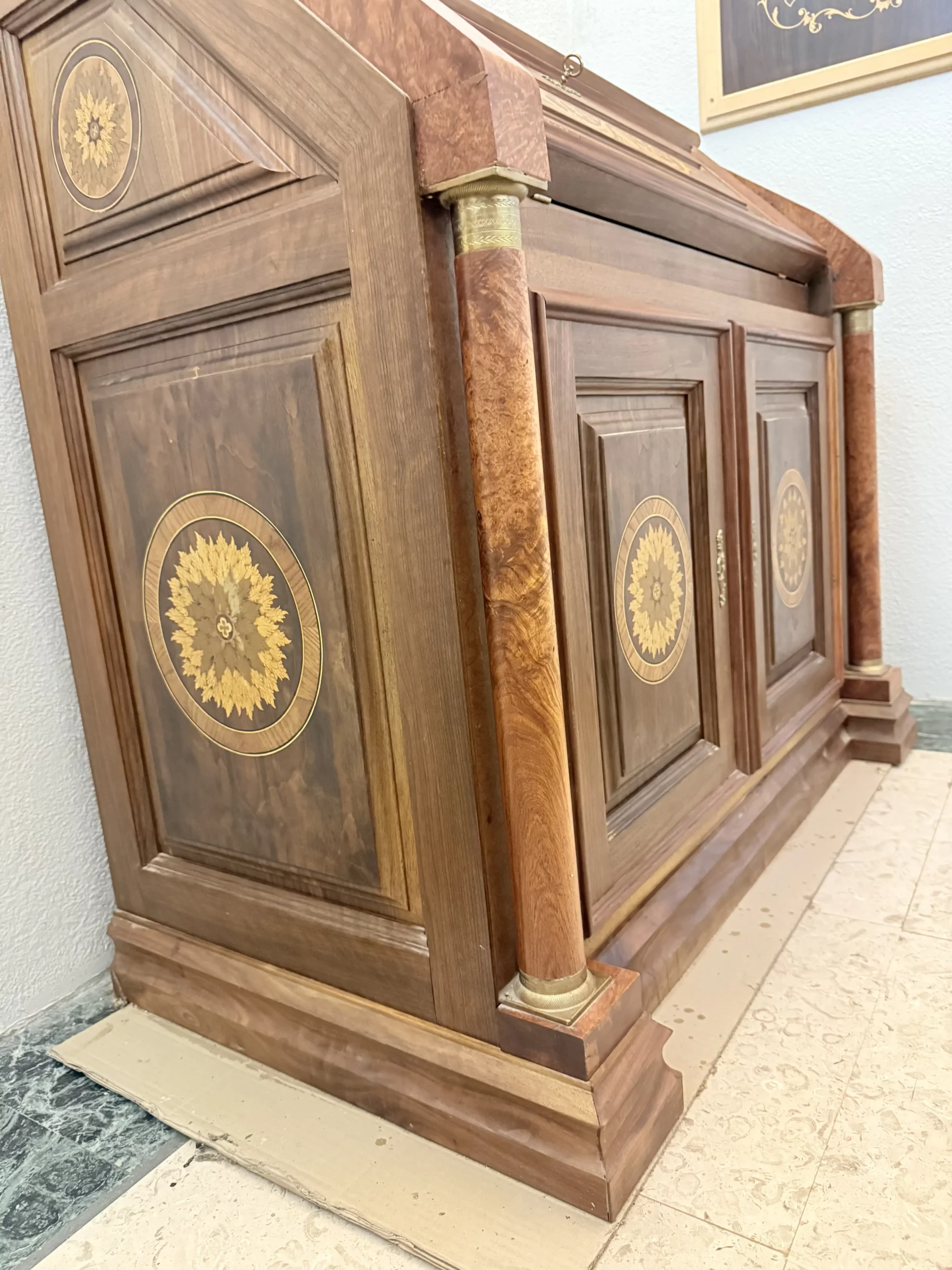 "Opera di Ay Mohsen realizzata con la tecnica dell'intarsio. Il titolo è Scrittoio delle ghirlande e rientra nella categoria Ebanisteria Monumentale. Lo Scrittoio delle Ghirlande di Aly Mohsen si configura come un mobile artistico intarsiato e scolpito, concepito come opera unica di alta ebanisteria, in cui funzione e articolazione decorativa sono integrate in una struttura di impianto architettonico. L’opera si colloca nella tradizione del mobile classico monumentale, reinterpretata attraverso una progettazione rigorosa e un controllo accurato dei materiali. La struttura portante è realizzata in noce calabrese della Sila, essenza selezionata per la compattezza, la stabilità strutturale e la profondità cromatica. I fianchi sagomati e le colonne frontali in radica di thuya introducono un ritmo plastico verticale, ulteriormente definito dalla presenza di elementi decorativi in bronzo che sottolineano i punti di raccordo e rafforzano la lettura architettonica del mobile. Elemento centrale dell’opera è il sistema di intarsi a ghirlanda, sviluppato secondo un disegno continuo e simmetrico. I profili degli intarsi sono eseguiti in acero, ebano e legno di rose, mentre i motivi floreali interni utilizzano una combinazione calibrata di acero, ebano verde, noce chiaro e noce scuro. La variazione cromatica tra le diverse essenze è costruita per gradazioni tonali e per equilibrio materico, in dialogo costante con il fondo ligneo. L’anta ribaltabile, incernierata sul fronte, rivela un interno organizzato con vani e cassetti in rovere, materiale scelto per la sua resistenza e per il contrasto visivo con le essenze esterne. Anche nella parte interna la lavorazione mantiene coerenza formale e qualità esecutiva, confermando l’unitarietà progettuale dell’opera. Il piano superiore e i bordi perimetrali sono trattati come una cornice strutturale e visiva, con funzione di contenimento dell’apparato decorativo e di definizione spaziale dell’insieme, contribuendo alla stabilità percettiva e alla chiarezza compositiva dello scrittoio."