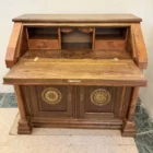 "Opera di Ay Mohsen realizzata con la tecnica dell'intarsio. Il titolo è Scrittoio delle ghirlande e rientra nella categoria Ebanisteria Monumentale. Lo Scrittoio delle Ghirlande di Aly Mohsen si configura come un mobile artistico intarsiato e scolpito, concepito come opera unica di alta ebanisteria, in cui funzione e articolazione decorativa sono integrate in una struttura di impianto architettonico. L’opera si colloca nella tradizione del mobile classico monumentale, reinterpretata attraverso una progettazione rigorosa e un controllo accurato dei materiali. La struttura portante è realizzata in noce calabrese della Sila, essenza selezionata per la compattezza, la stabilità strutturale e la profondità cromatica. I fianchi sagomati e le colonne frontali in radica di thuya introducono un ritmo plastico verticale, ulteriormente definito dalla presenza di elementi decorativi in bronzo che sottolineano i punti di raccordo e rafforzano la lettura architettonica del mobile. Elemento centrale dell’opera è il sistema di intarsi a ghirlanda, sviluppato secondo un disegno continuo e simmetrico. I profili degli intarsi sono eseguiti in acero, ebano e legno di rose, mentre i motivi floreali interni utilizzano una combinazione calibrata di acero, ebano verde, noce chiaro e noce scuro. La variazione cromatica tra le diverse essenze è costruita per gradazioni tonali e per equilibrio materico, in dialogo costante con il fondo ligneo. L’anta ribaltabile, incernierata sul fronte, rivela un interno organizzato con vani e cassetti in rovere, materiale scelto per la sua resistenza e per il contrasto visivo con le essenze esterne. Anche nella parte interna la lavorazione mantiene coerenza formale e qualità esecutiva, confermando l’unitarietà progettuale dell’opera. Il piano superiore e i bordi perimetrali sono trattati come una cornice strutturale e visiva, con funzione di contenimento dell’apparato decorativo e di definizione spaziale dell’insieme, contribuendo alla stabilità percettiva e alla chiarezza compositiva dello scrittoio."